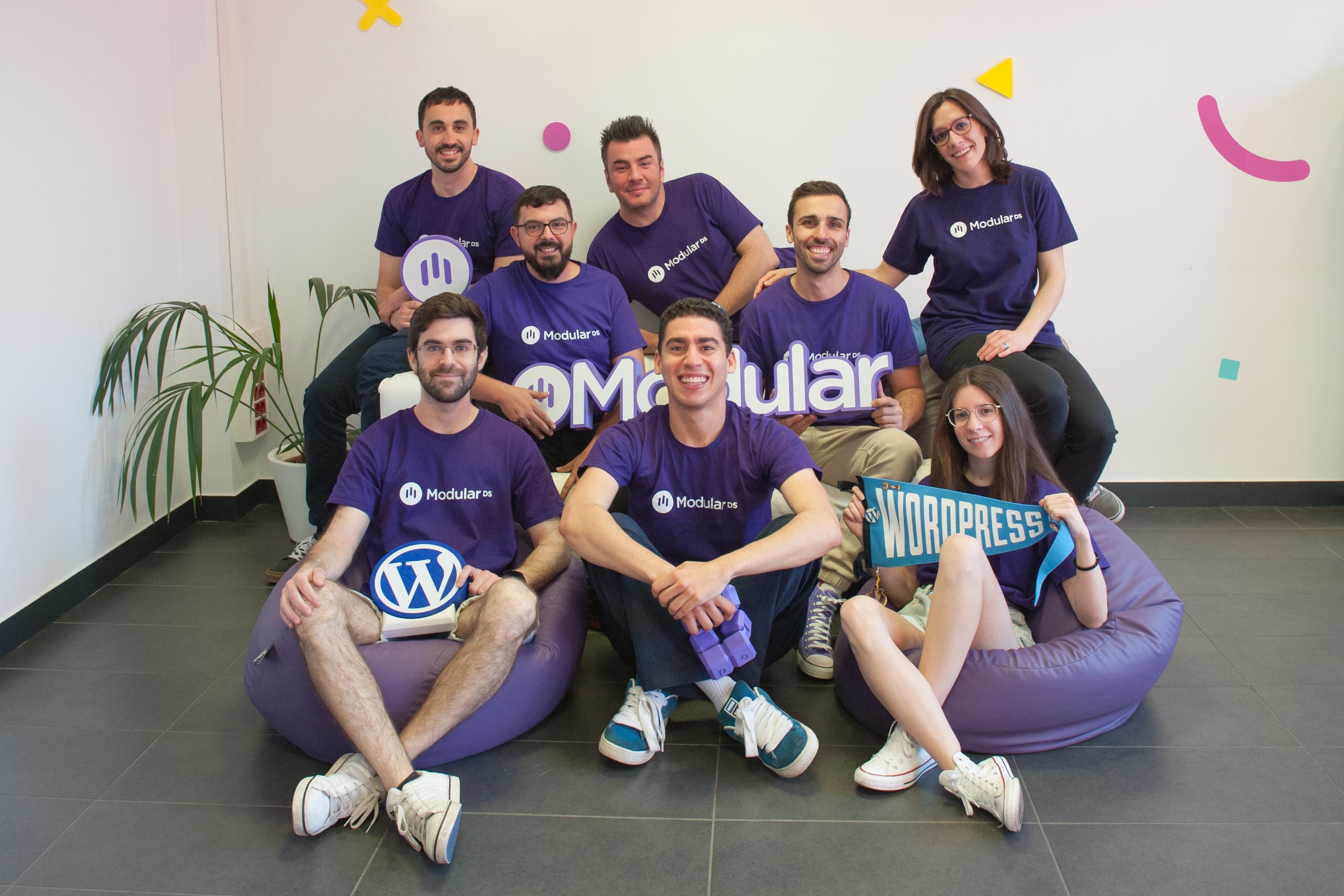 The Modular DS team together in the office, wearing purple Modular DS t-shirts, with the WordPress logo and a WordPress pennant.